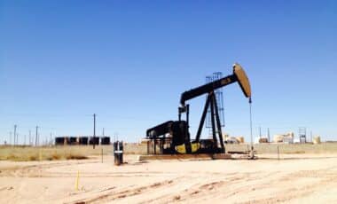 Cette image montre une pompe à balancier (derrick) utilisée pour l'extraction du pétrole, installée dans une zone désertique avec un ciel dégagé. En arrière-plan, on distingue des structures industrielles et des réservoirs.