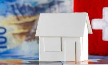 maison en papier blanc sur fond de billets en francs suisses et drapeau suisse