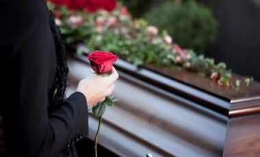 Photo d'un cercueil en bois dans un cimetière en face duquel se tient une personne tenant une rose rouge dans sa main droite.