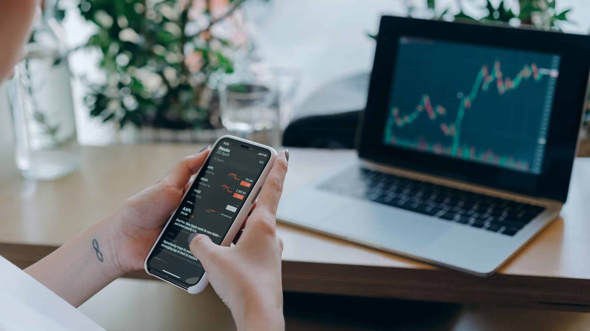 Personne utilisant un smartphone et un ordinateur portable pour analyser des graphiques financiers et suivre ses investissements.