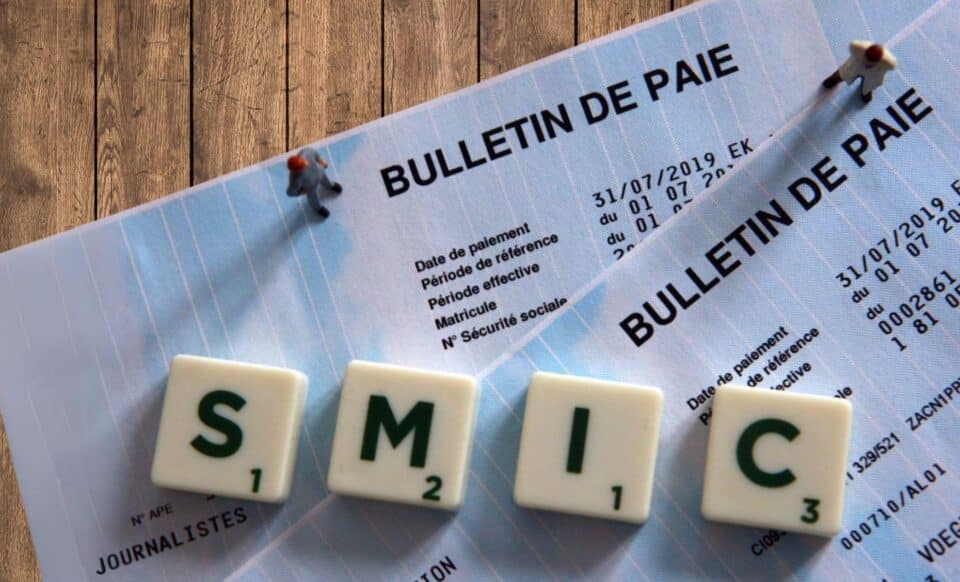 Photo de tuiles formant l'acronyme « Smic » par-dessus deux bulletins de paie posés sur un support en bois