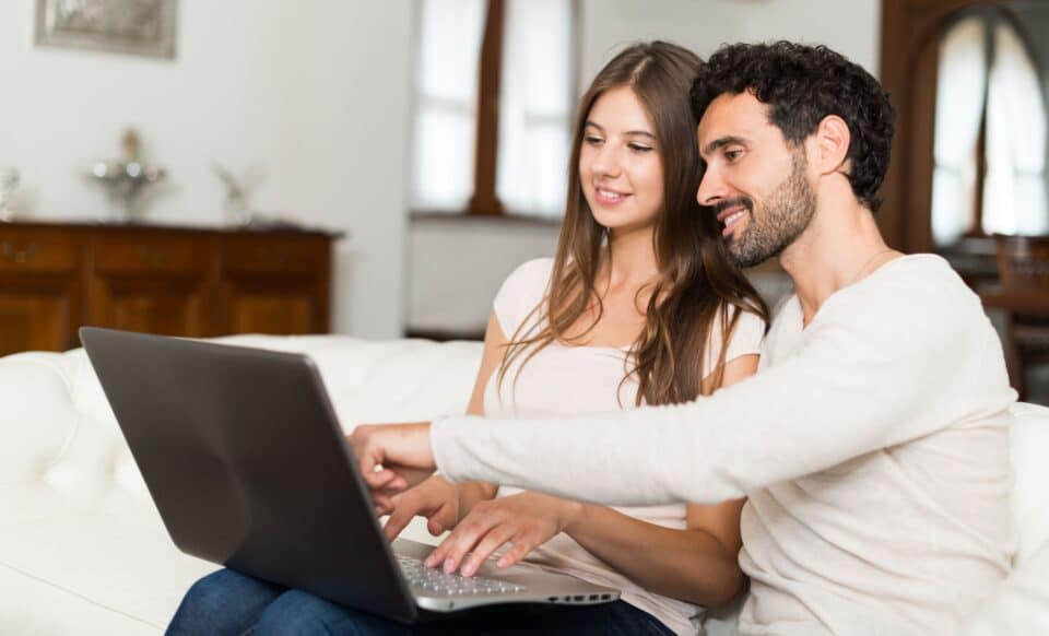 Un couple assis sur un canapé utilisant un ordinateur portable, discutant et consultant des informations, dans une ambiance détendue à domicile