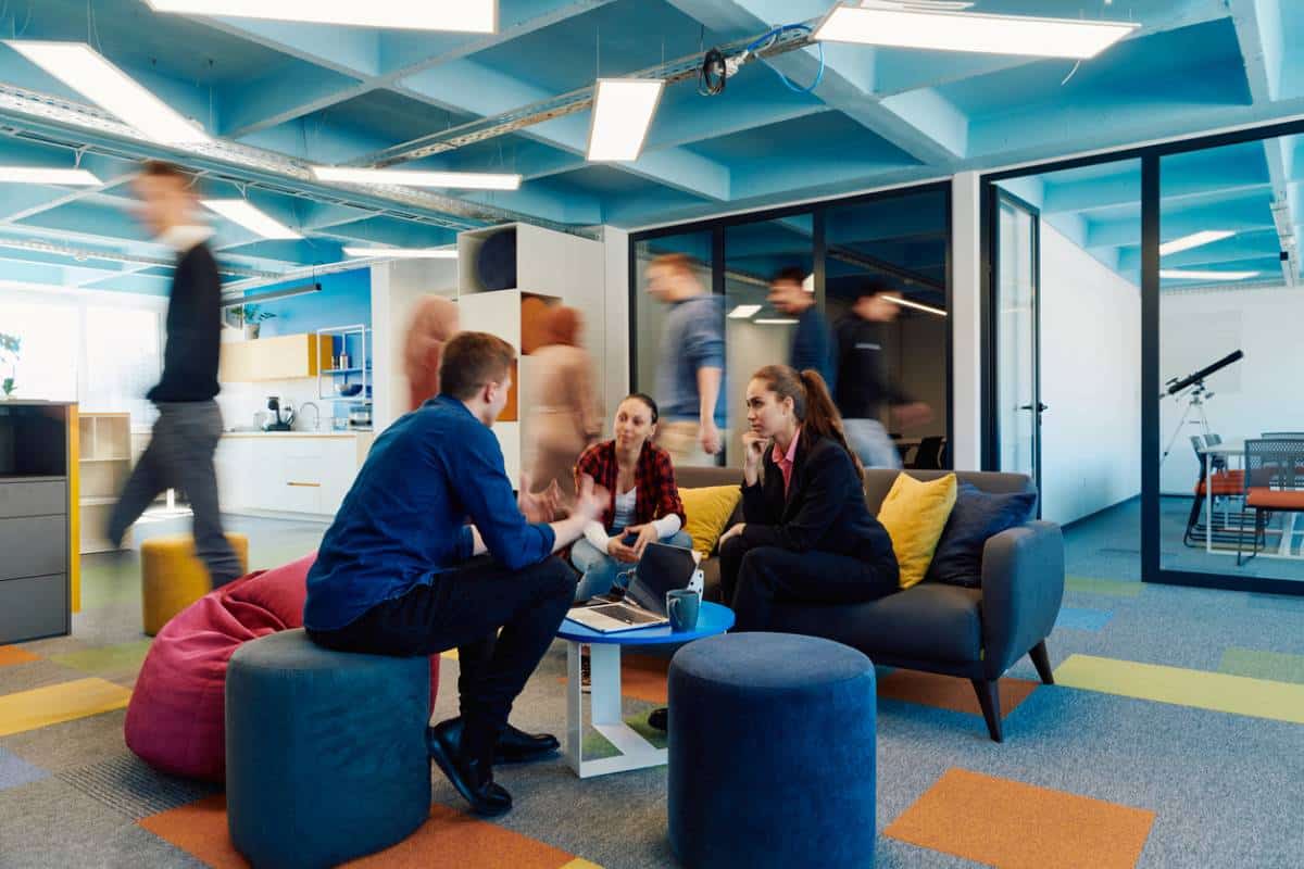 Espace de travail collaboratif avec des employés discutant dans un salon moderne, entouré de mobilier coloré et de poufs, avec des personnes en mouvement en arrière-plan dans un bureau à aire ouverte.