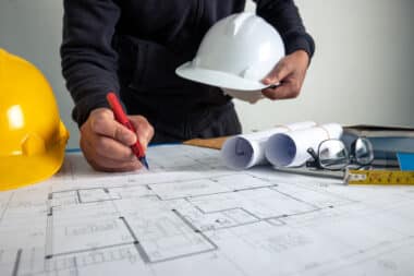 Personne travaillant sur des plans architecturaux avec un casque blanc dans une main et un stylo rouge dans l'autre, entourée d'un casque jaune, de plans roulés, de lunettes et d'un ruban à mesurer sur la table. Environnement de travail lié à la construction ou à l'architecture.