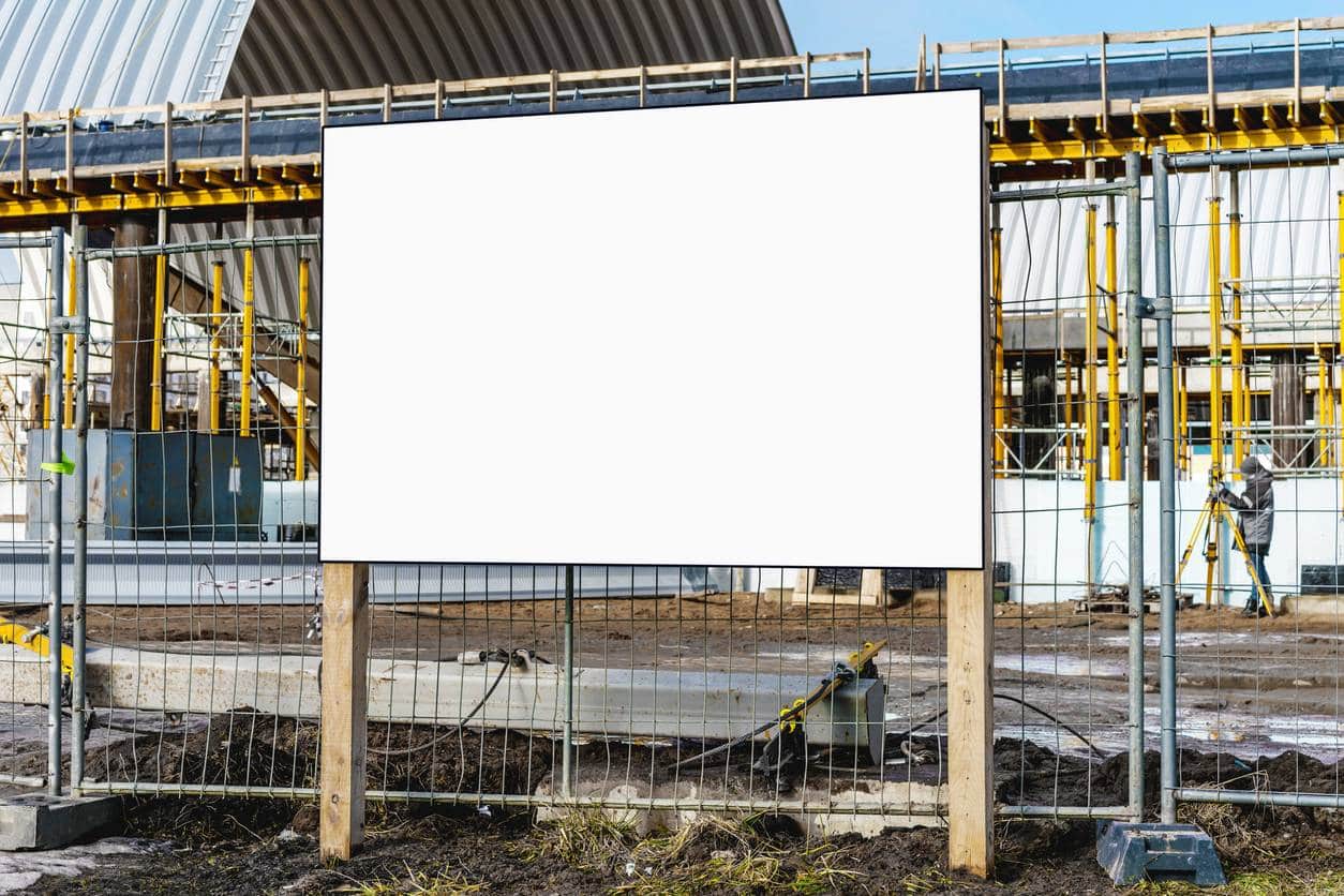 Panneau d'affichage blanc sur un chantier de construction, fixé à une clôture métallique avec des poteaux en bois, entouré de structures de construction et d'ouvriers en activité.