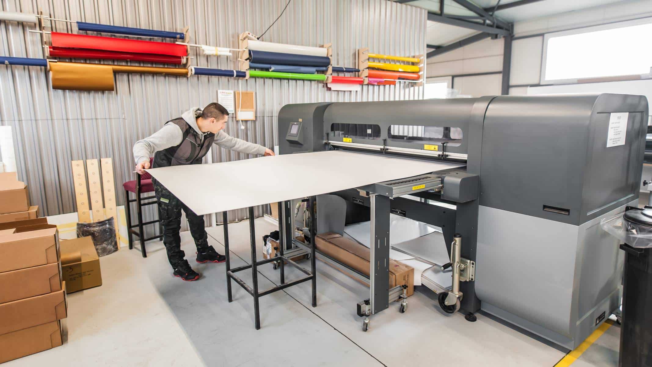 Opérateur utilisant une grande imprimante industrielle dans un atelier, manipulant une feuille de matériau, entouré de rouleaux de matériaux colorés et de boîtes en carton. Environnement organisé pour la production à grande échelle.