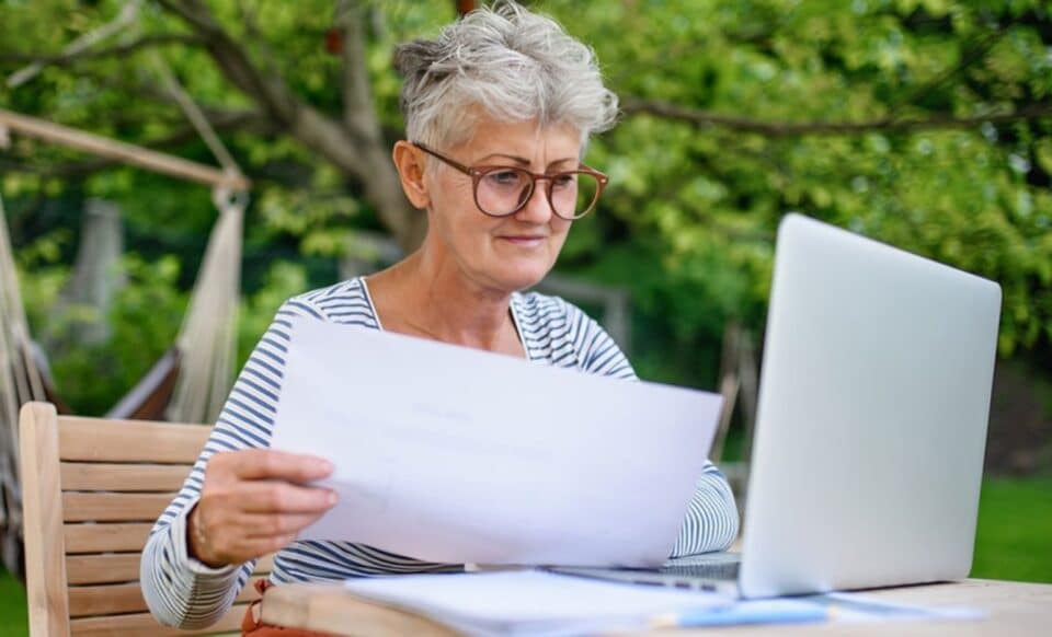 Une femme âgée assise à une table en extérieur, tenant des documents et regardant son ordinateur portable pour illustrer la retraite