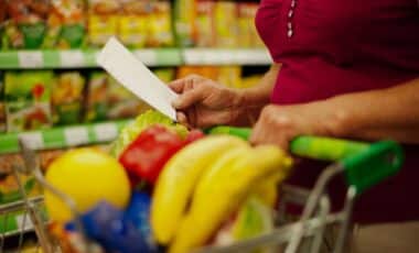 Photo d'une consommatrice consultant sa liste de courses avec un charriot de supermarché plein de denrées pour illustrer le sujet de l'inflation