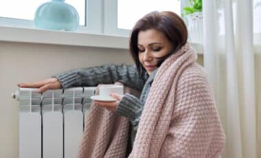 Tasse à la main et couverte d'un plaid, une femme qui a visiblement froid se chauffe tout près de son radiateur