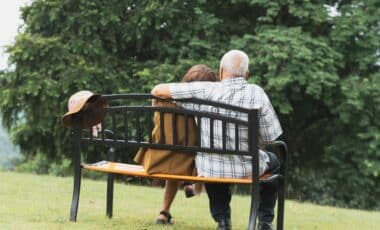 Cette image montre un couple de seniors assis sur un banc dans un parc, symbolisant un moment de sérénité et de détente, souvent associé à la retraite. Le cadre naturel en arrière-plan renforce l'idée de repos et de qualité de vie après une carrière active.