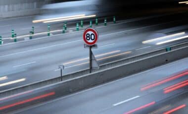 Cette image montre un panneau de limitation de vitesse à 80 km/h sur une autoroute, entouré de véhicules en mouvement rapide. Elle illustre les règles de circulation et les ajustements auxquels les automobilistes doivent se conformer, en particulier dans le cadre de nouvelles mesures visant à renforcer la sécurité routière et réduire les émissions polluantes.