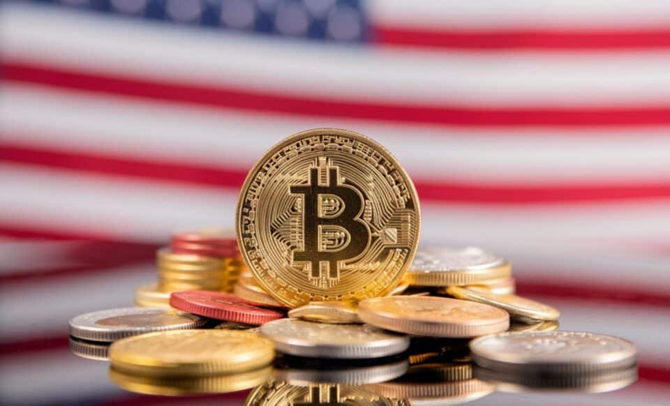 Une pièce de bitcoin en or placé sur plusieurs autres pièces, avec en arrière plan le drapeau des Etats-Unis