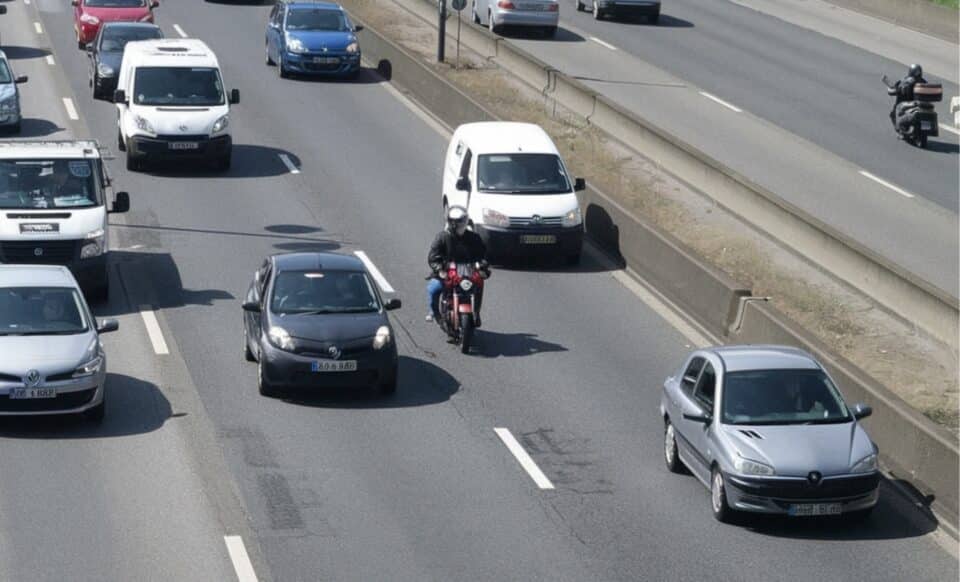 L'image montre un motard circulant entre des files de voitures sur une route à plusieurs voies, illustrant la pratique de la circulation interfiles.