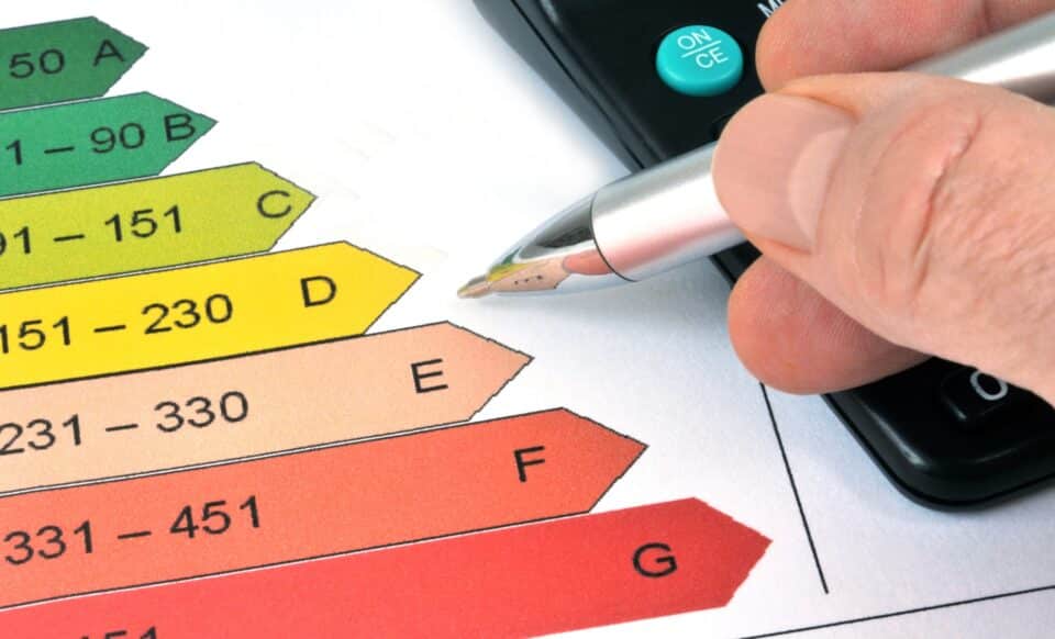 L'image montre une échelle de performance énergétique typique d’un Diagnostic de performance énergétique (DPE), avec les classes allant de A (vert) à G (rouge). Une main pointe avec un stylo.