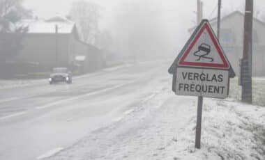 Cette image montre une route partiellement enneigée et verglacée, avec un panneau de signalisation indiquant "Verglas fréquent". Une voiture circule prudemment sous un ciel gris et brumeux, illustrant des conditions météo hivernales typiques.