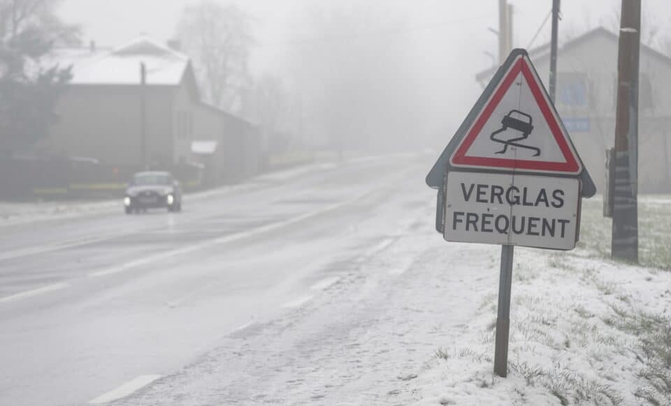 Cette image montre une route partiellement enneigée et verglacée, avec un panneau de signalisation indiquant "Verglas fréquent". Une voiture circule prudemment sous un ciel gris et brumeux, illustrant des conditions météo hivernales typiques.