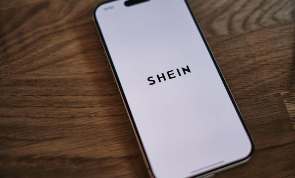 L'image montre un smartphone affichant le logo de Shein sur un écran blanc, posé sur une surface en bois.