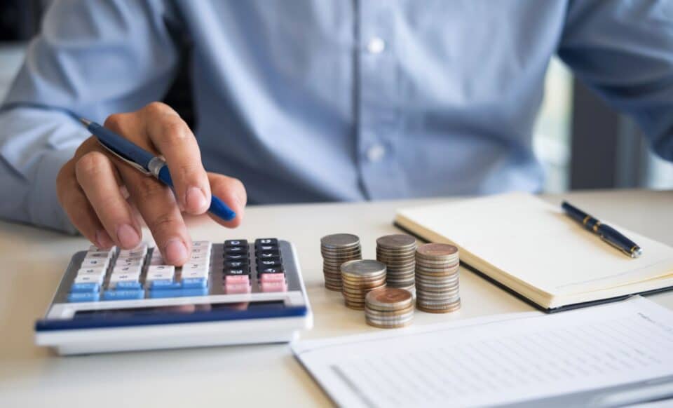 une personne assise sur un bureau qui utilise une calculatrice devant des tas de pièces de monnaie pour évoquer la baisse du taux du PEL