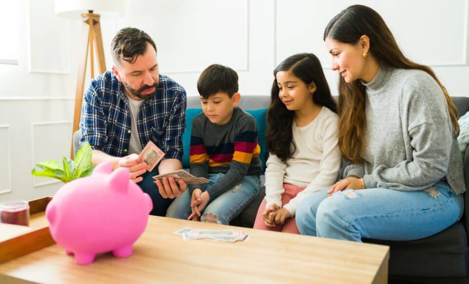 Cette image montre une famille réunie autour d'une table, avec un père comptant de l'argent sous le regard attentif de ses enfants et de leur mère. Elle illustre le rôle des allocations familiales dans le soutien financier des familles, mettant en lumière l'importance de ces aides pour la gestion des dépenses du foyer.