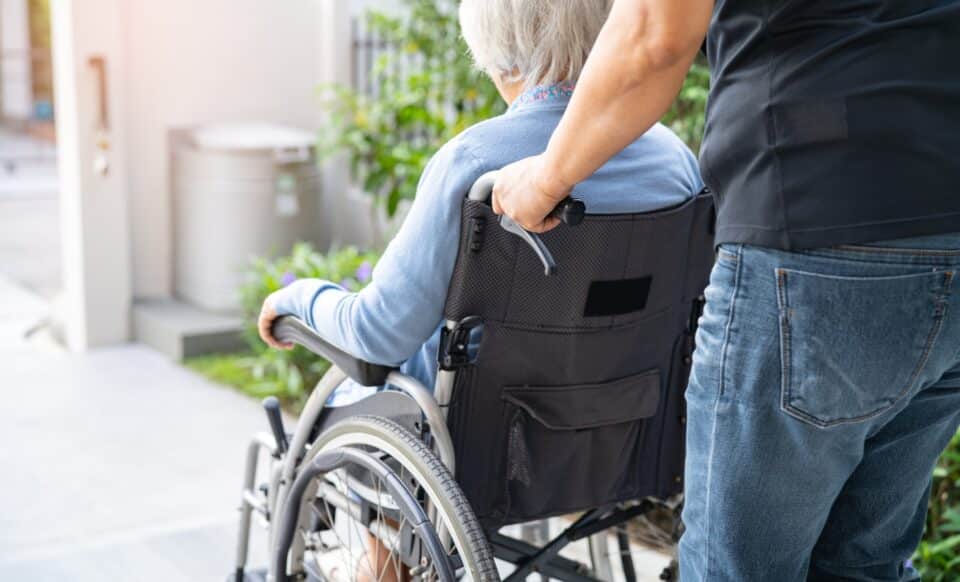 L'image montre une personne âgée en fauteuil roulant, assistée par un aidant, dans un environnement extérieur. Elle illustre le soutien nécessaire pour les personnes en situation de dépendance, un contexte lié à l'éligibilité à l'Aspa pour garantir une aide financière aux retraités aux faibles ressources.