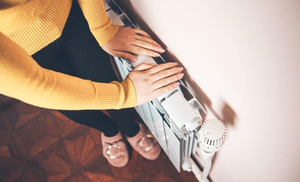 L'image montre une personne en pull jaune posant ses mains sur un radiateur, évoquant le sujet du chauffage domestique et de la gestion de la température.