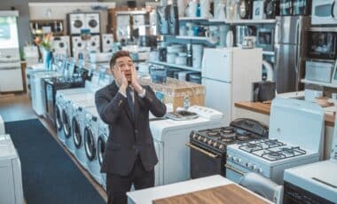 L'image montre un homme en costume dans un magasin d'électroménager, visiblement désorienté ou frustré devant une rangée d'appareils tels que des lave-linges et des réfrigérateurs.