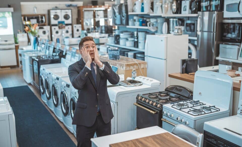 L'image montre un homme en costume dans un magasin d'électroménager, visiblement désorienté ou frustré devant une rangée d'appareils tels que des lave-linges et des réfrigérateurs.