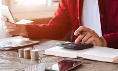 L'image montre une personne en train de gérer ses finances personnelles. Elle utilise une calculatrice tout en consultant un carnet et des documents. Sur la table, on distingue des piles de pièces de monnaie, symbolisant l'épargne, et un smartphone à proximité.