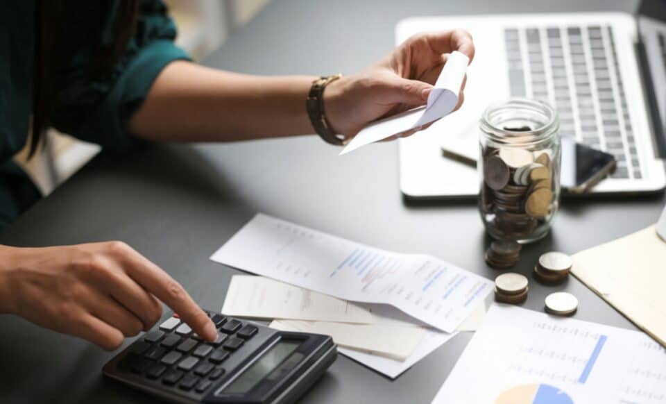 L'image montre une personne en train de gérer ses finances à l'aide d'une calculatrice, de reçus, et d'un ordinateur portable. Un bocal rempli de pièces de monnaie est visible, symbolisant une planification budgétaire minutieuse, un thème pertinent pour les hausses attendues en janvier 2025.