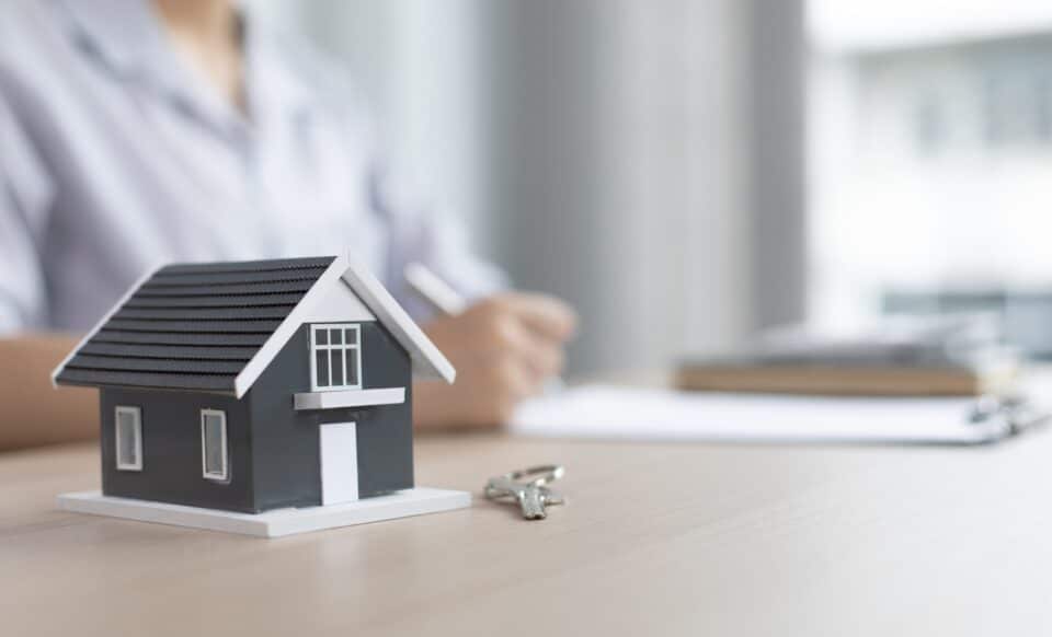 Cette image montre une maquette de maison posée sur une table, accompagnée d’un trousseau de clés. En arrière-plan, une personne écrit sur des documents, symbolisant la gestion administrative ou la demande liée au logement social.