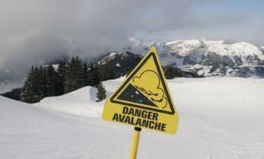 Cette image montre un paysage montagneux enneigé, avec un panneau jaune indiquant un danger d'avalanche. Le ciel est partiellement couvert, reflétant des conditions météo potentiellement instables, typiques des zones sujettes aux avalanches en période hivernale.