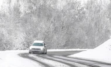 L'image montre une voiture circulant sur une route enneigée entourée d'arbres givrés, créant un paysage hivernal typique. Cette scène évoque des conditions météo difficiles, avec des routes glissantes et un environnement marqué par la neige, soulignant la prudence nécessaire lors de déplacements dans de telles conditions.