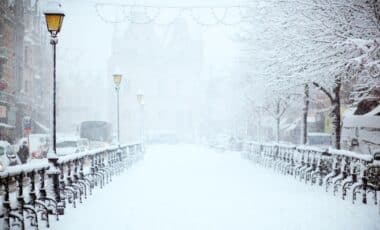 Cette image illustre un paysage urbain recouvert d'une épaisse couche de neige, avec des lampadaires et des arbres givrés bordant une rue. Elle évoque une météo hivernale typique, marquée par des chutes de neige abondantes, des températures froides et une ambiance paisible mais glaciale.