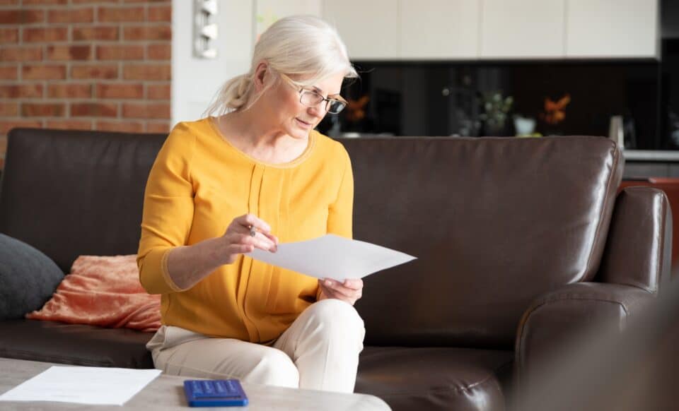 Cette image montre une femme âgée, assise sur un canapé, examinant un document, symbolisant des démarches administratives liées à la pension de réversion.