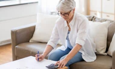 Cette image montre une senior assise sur un canapé, concentrée sur des documents et utilisant une calculatrice pour illustrer la pension de réversion.