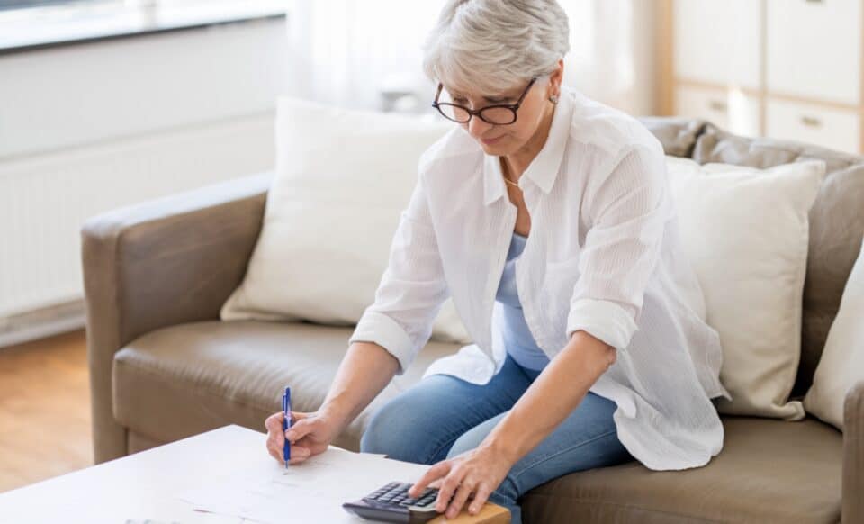 Cette image montre une senior assise sur un canapé, concentrée sur des documents et utilisant une calculatrice pour illustrer la pension de réversion.