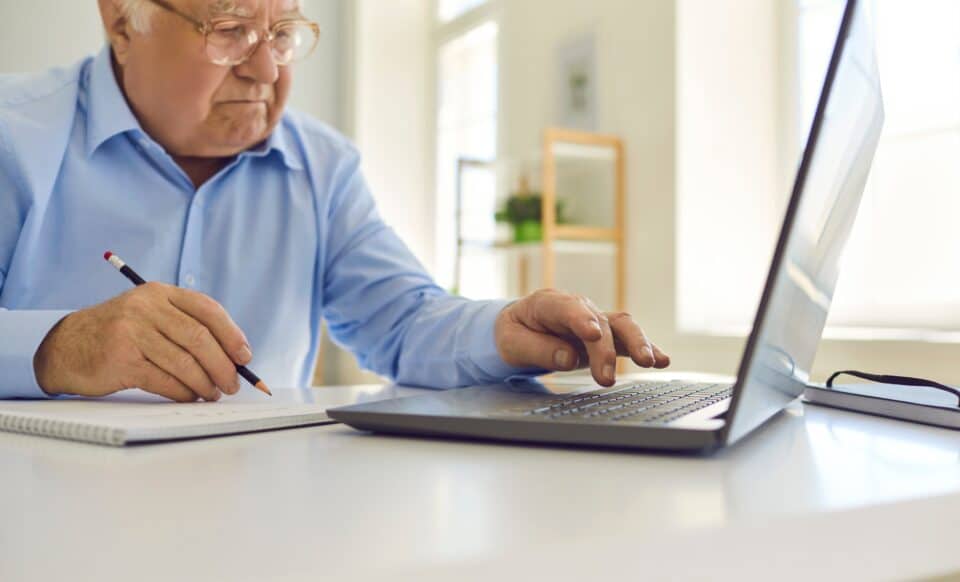 Cette image montre un homme âgé concentré sur son travail, prenant des notes tout en utilisant un ordinateur portable. Elle illustre la réflexion et la planification, symbolisant des démarches telles que la préparation de la retraite ou la gestion des finances personnelles.