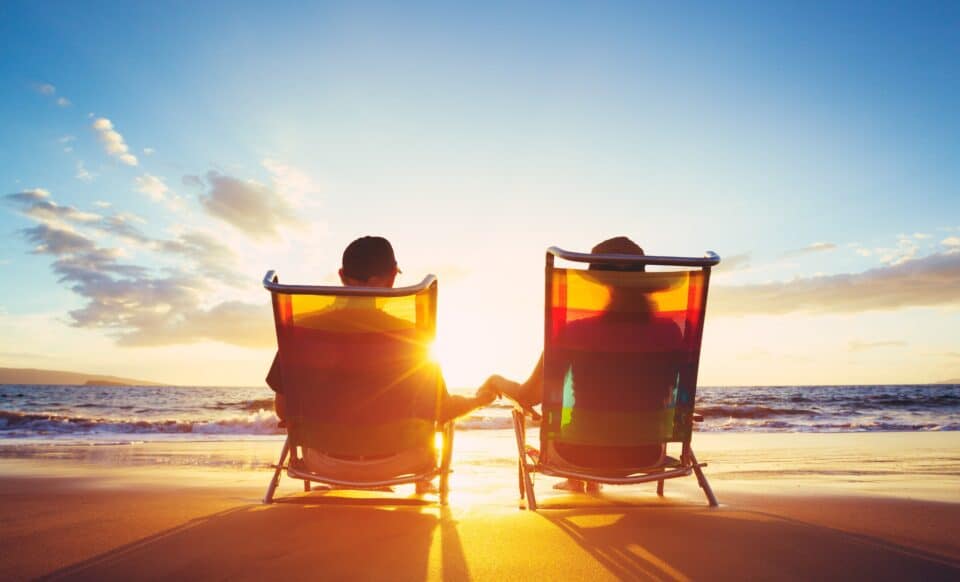 Cette image montre un couple assis sur des chaises longues, face à la mer au coucher du soleil. Elle symbolise l'idée d'une retraite paisible et sereine à l'étranger, dans un cadre idyllique et ensoleillé.