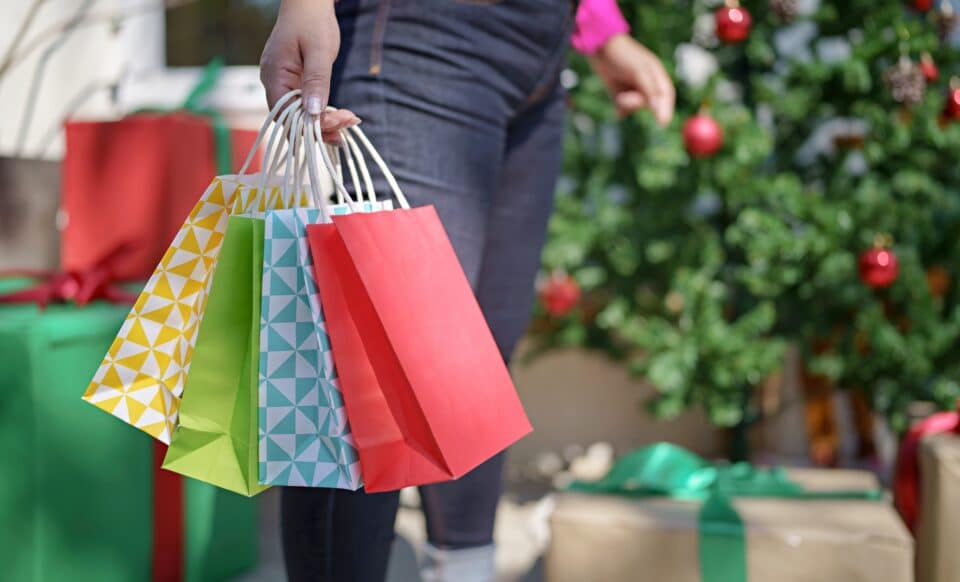 Cette image montre une personne tenant plusieurs sacs colorés devant un décor festif, évoquant la période des soldes d'hiver. Les décorations de Noël en arrière-plan renforcent l'idée des achats de fin d'année ou du début d'année, souvent marqués par des promotions attractives.