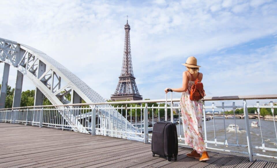 L'image illustre une scène typique de tourisme à Paris, avec une femme vêtue d'une robe estivale, un chapeau, et un sac à dos, accompagnée d'une valise. Elle se tient sur un pont métallique, face à la Tour Eiffel, symbole emblématique de la ville. L'environnement suggère une journée ensoleillée, propice à la découverte et aux voyages.