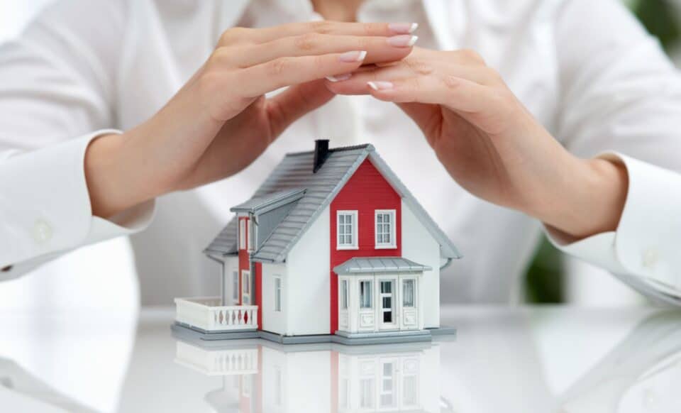 une personne, probablement une femme, aux doigts manucurés, qui couvre une maison miniature avec ses mains pour évoquer l'assurance habitation