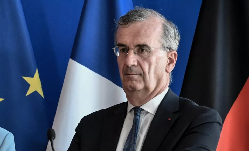 L'image montre François Villeroy de Galhau, gouverneur de la Banque de France, devant les drapeaux français et européen, évoquant un discours officiel sur le déficit public et la maîtrise des finances.