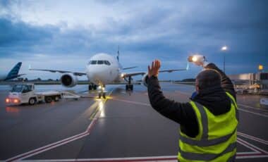 un grand avion de couleur blanche sur le tarmac d'un aéroport, où se tient un membre de l'équipage qui effectue un signalement muni d'une lampe torche pour évoquer la surtaxe sur les billets d'avion