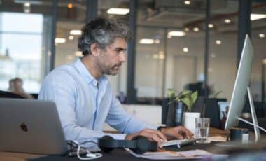 Un homme aux cheveux gris vetue d'une chemise bleu, assis devant un ordinateur dans un bureau moderne pour évoquer le travail en freelance.