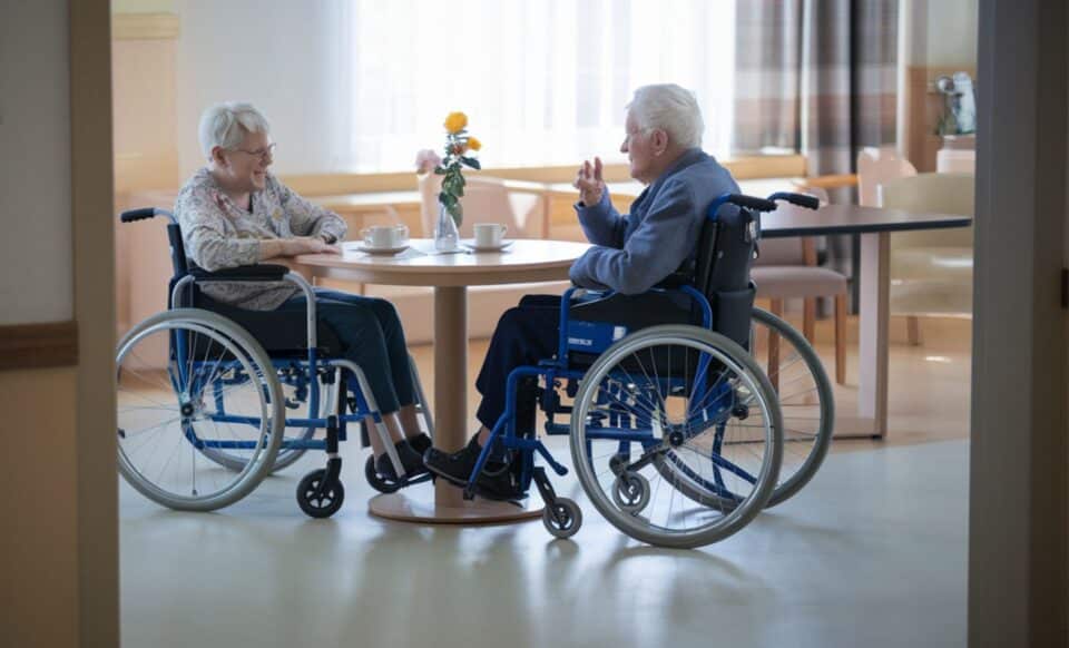 deux personnes agées en fauteuil roulant assises autour d'une table sur laquelle se trouve un vase pour évoquer les EHPAD