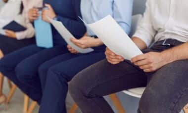 Des candidats assis en file d’attente tiennent des documents, illustrant une session de recrutement ou un entretien d’emploi.