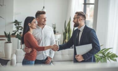 Un homme en costume, incarnant un agent immobilier, serre la main d'une femme vêtue d'un T-shirt orange aux côtés d'un homme vêtu d'une chemise blanche, incarnant des acheteurs