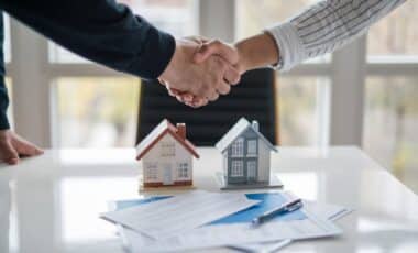 deux individus qui se serrent la main sur un bureau sur lequel se trouve deux petite maisons et des contrats