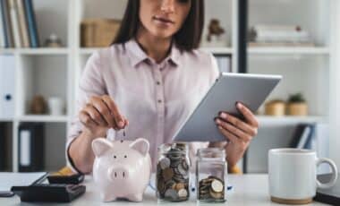 Une jeune personne avec une tablette, glissant une pièce dans une tirelire. à côté, un bocal contenant des pièces de monnaie, le tout pour illustrer le Livret jeune