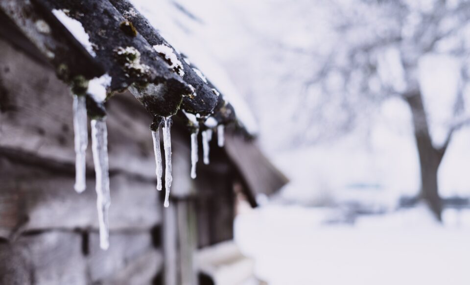 L'image montre des stalactites de glace suspendues au bord d'un toit en bois, avec un paysage enneigé flou en arrière-plan. Cette scène hivernale illustre des conditions météo marqué par un froid intense, typiques des épisodes de neige et verglas.